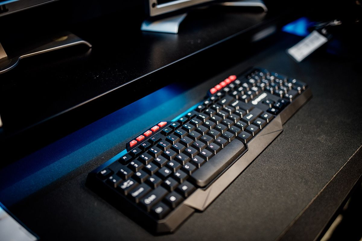 A computer keyboard is sitting on a dark desk in an office setup. The keyboard has a mix of black and red keys. Background shows some monito
