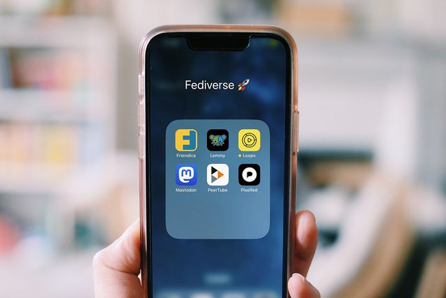 a photo of a hand holding a smartphone; on screen you see icons of Fediverse apps: Lemmy, Loops, Mastodon, PeerTube and 