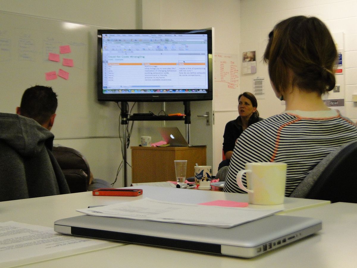 Students in front of large screen. In the foreground a closed laptop. Most likely qualitative data analysis in a spreadsheet, but it could a