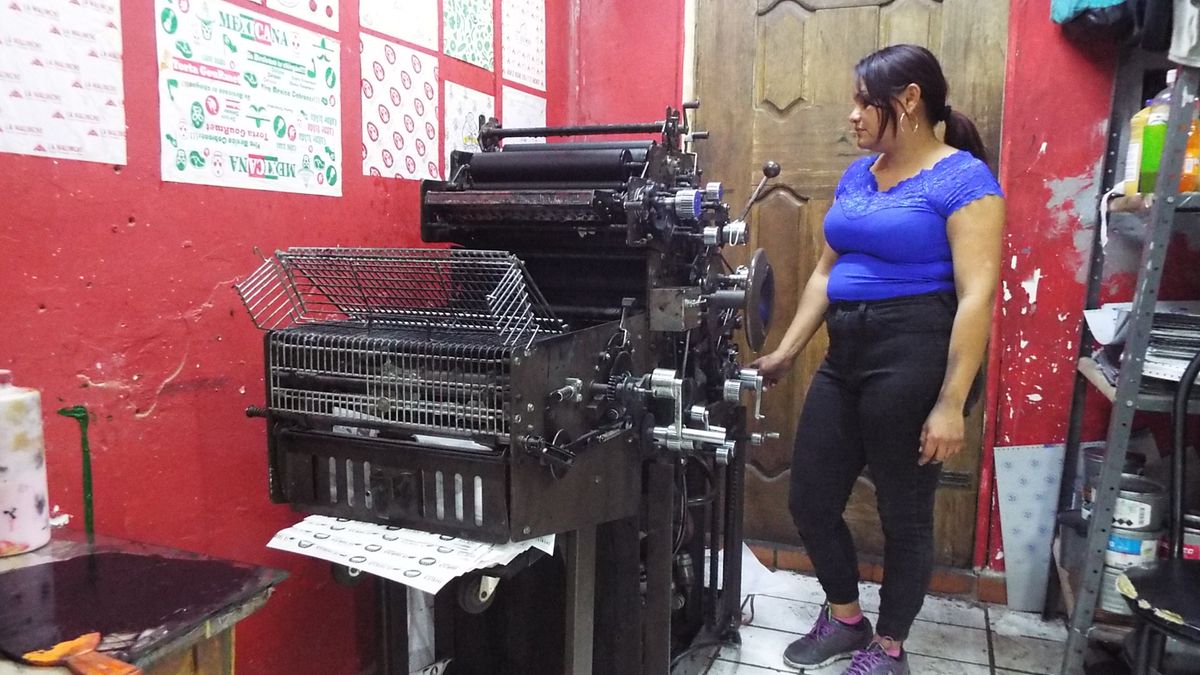 Mujer de color azul vigila una maquina de impresión offset en un cuarto pintado de rojo con diversas hojas impresas pegadas en la pared.